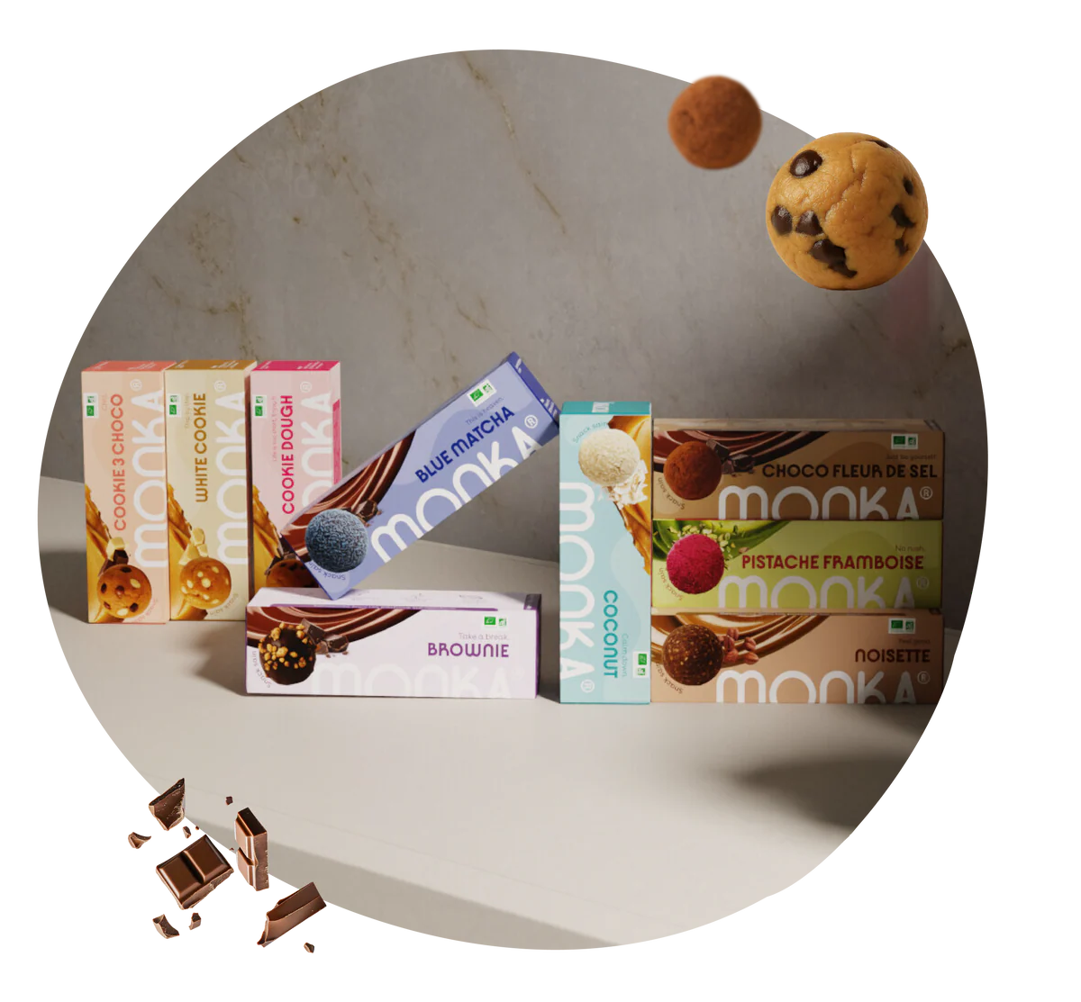 Différents paquets de snacks Monka de saveurs variées comme cookie, brownie, matcha bleu, noix de coco, chocolat fleur de sel, pistache framboise et noisette, avec des boules de snack flottantes et des morceaux de chocolat sur une table.