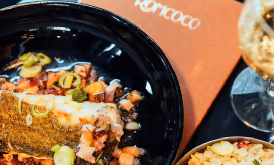 Plat de poisson grillé garni de dés de légumes et herbes dans un bol noir, accompagné d'un verre et d'un bol de céréales.