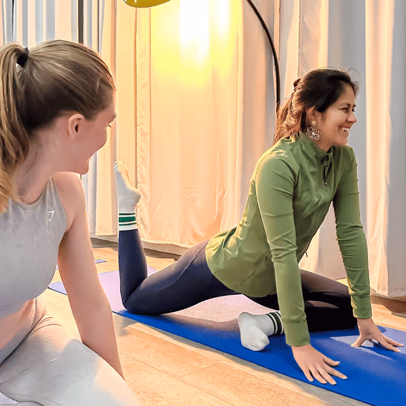 Deux femmes souriantes en tenue de sport, l'une effectue un étirement au sol sur un tapis bleu, l'autre l'encourage en face d'elle.