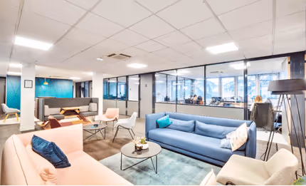 Espace de détente moderne avec canapés colorés, tables basses, et bureaux visibles à travers des cloisons vitrées.