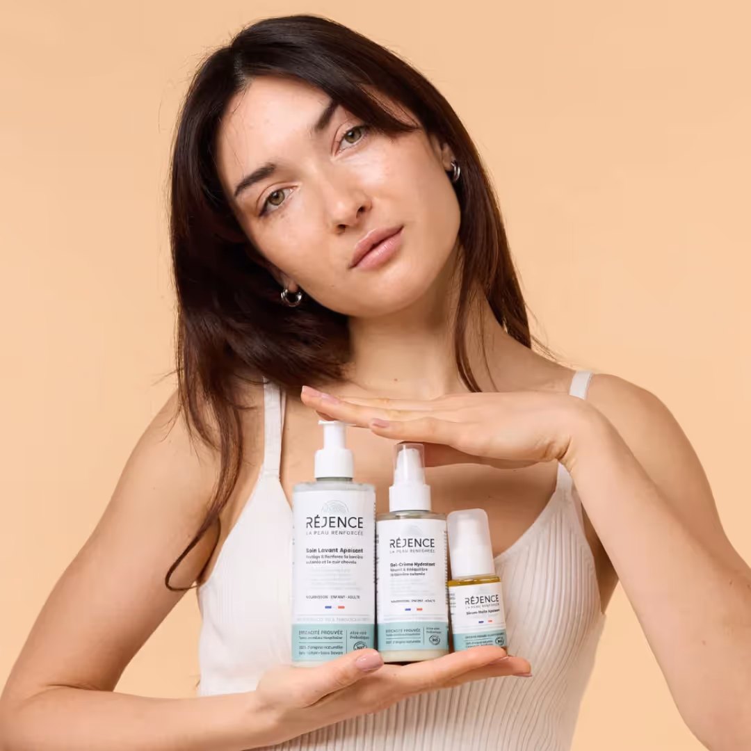 Une femme tenant trois produits de soin Réjence devant un fond beige clair.