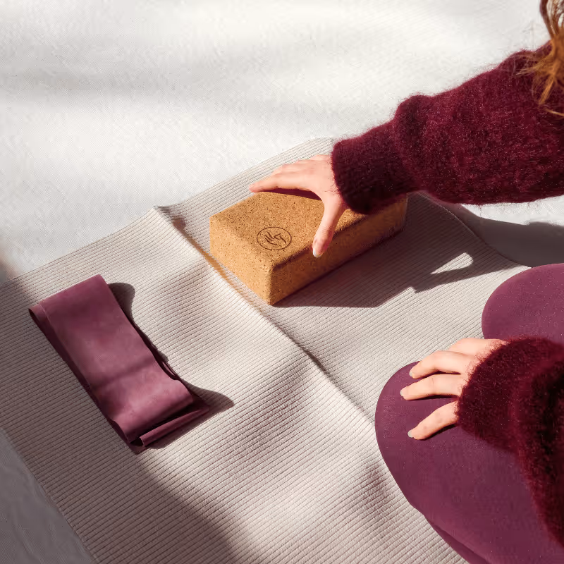 Person agenouillée sur un tapis de yoga beige, tenant un bloc de yoga en liège à côté d'une bande de résistance violette.