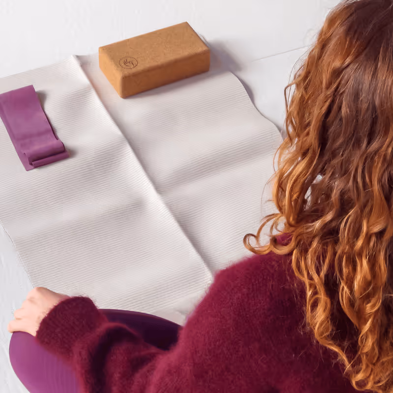 Personne cheveux roux assise en position yoga sur un tapis beige avec une sangle violette et un bloc en liège.