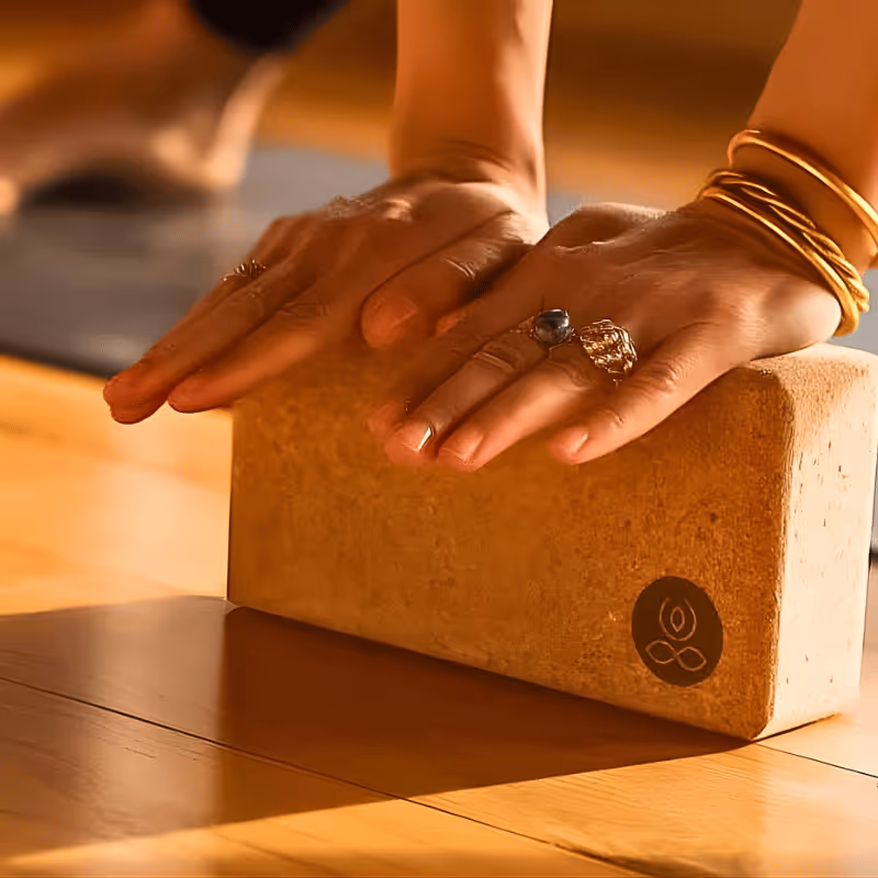 Mains posées sur un bloc de yoga en liège sur un sol en bois avec bracelets dorés et bagues.