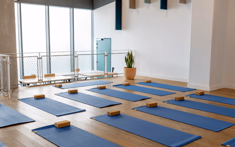 Salle de yoga avec plusieurs tapis bleus alignés au sol, chacun équipé d'un ballon bleu, deux briques en bois et une sangle rouge.