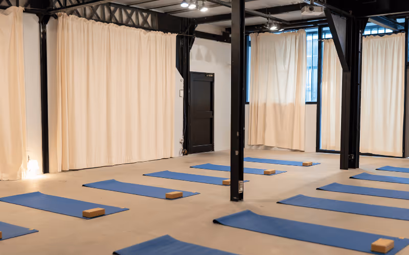 Salle de yoga avec plusieurs tapis bleus alignés au sol, chacun équipé d'un ballon bleu, deux briques en bois et une sangle rouge.