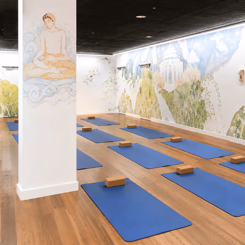 Salle de yoga avec tapis bleus et briques, murs décorés de fresques et colonne avec illustration d'une figure méditative.