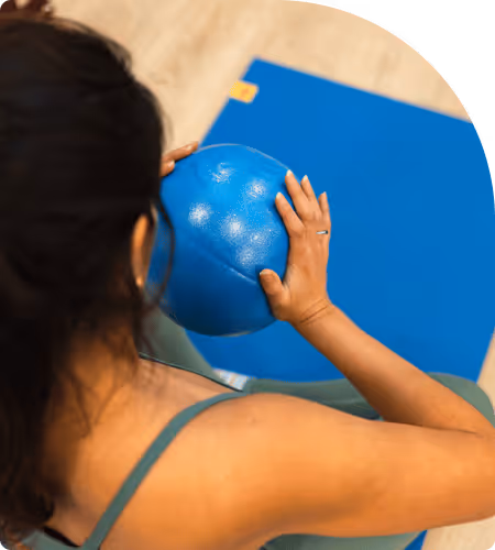 Femme vue de dos tenant un ballon de fitness bleu sur un tapis de yoga bleu.