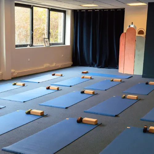 Une femme s'exerçant en posture de yoga Guerrier II devant un miroir dans une salle lumineuse avec un tapis bleu.