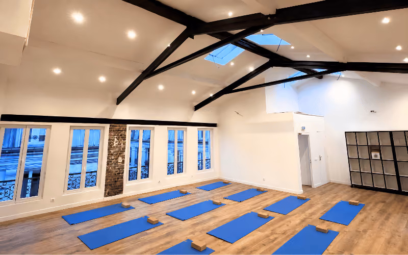 Salle du studio Yoze Ménilmontant 2 avec tapis bleus, blocs de yoga et poutres apparentes.