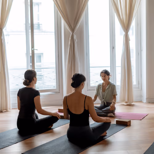 Une femme s'exerçant en posture de yoga Guerrier II devant un miroir dans une salle lumineuse avec un tapis bleu.