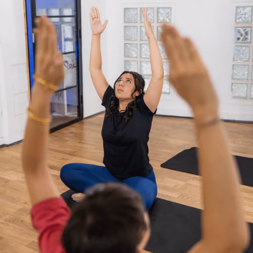 Une femme s'exerçant en posture de yoga Guerrier II devant un miroir dans une salle lumineuse avec un tapis bleu.