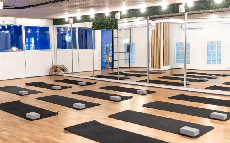 Salle de yoga avec plusieurs tapis bleus alignés au sol, chacun équipé d'un ballon bleu, deux briques en bois et une sangle rouge.