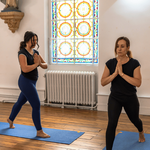 Une femme s'exerçant en posture de yoga Guerrier II devant un miroir dans une salle lumineuse avec un tapis bleu.
