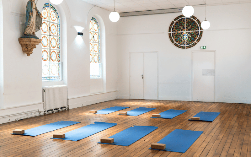 Salle de yoga avec plusieurs tapis bleus alignés au sol, chacun équipé d'un ballon bleu, deux briques en bois et une sangle rouge.