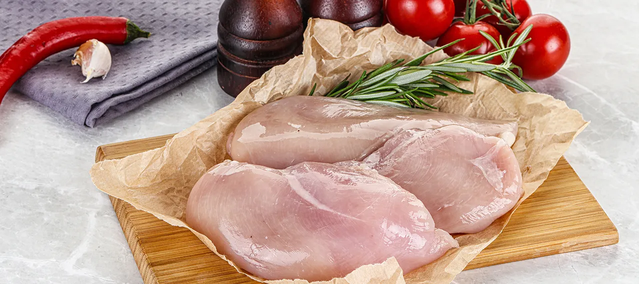 Two raw chicken breasts on parchment paper and a wooden cutting board with rosemary, cherry tomatoes, chili pepper, and garlic clove nearby.