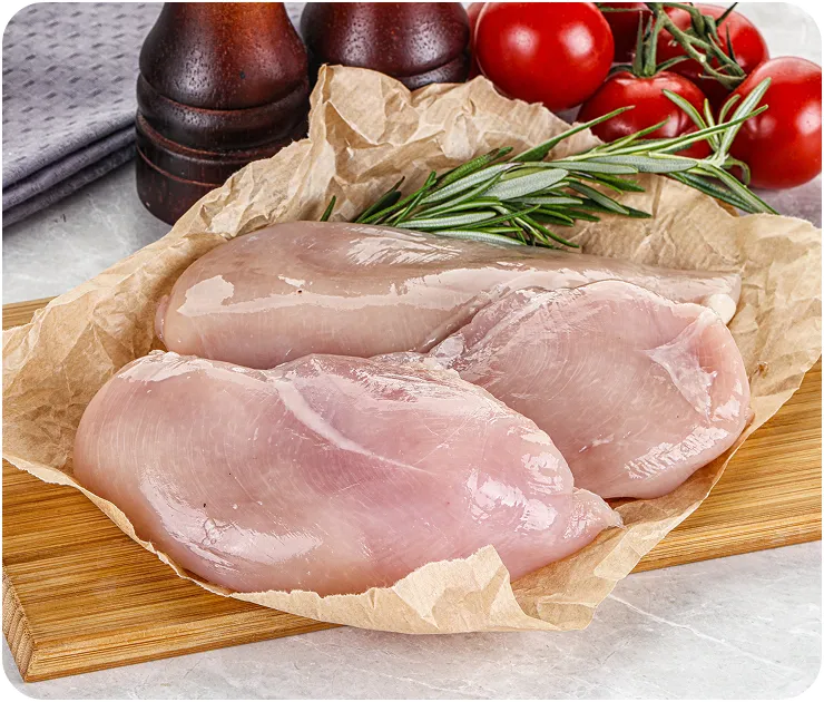 Two raw chicken breasts on parchment paper and a wooden cutting board with rosemary, cherry tomatoes, chili pepper, and garlic clove nearby.