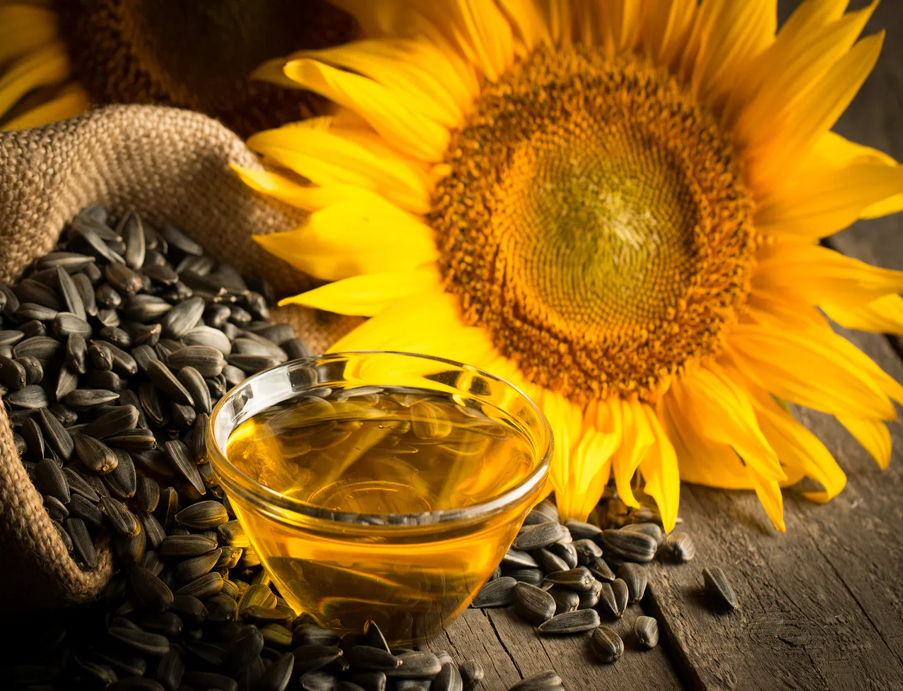 Glass bowl of sunflower oil surrounded by sunflower seeds spilling from a burlap sack and a large bright sunflower on a wooden surface.