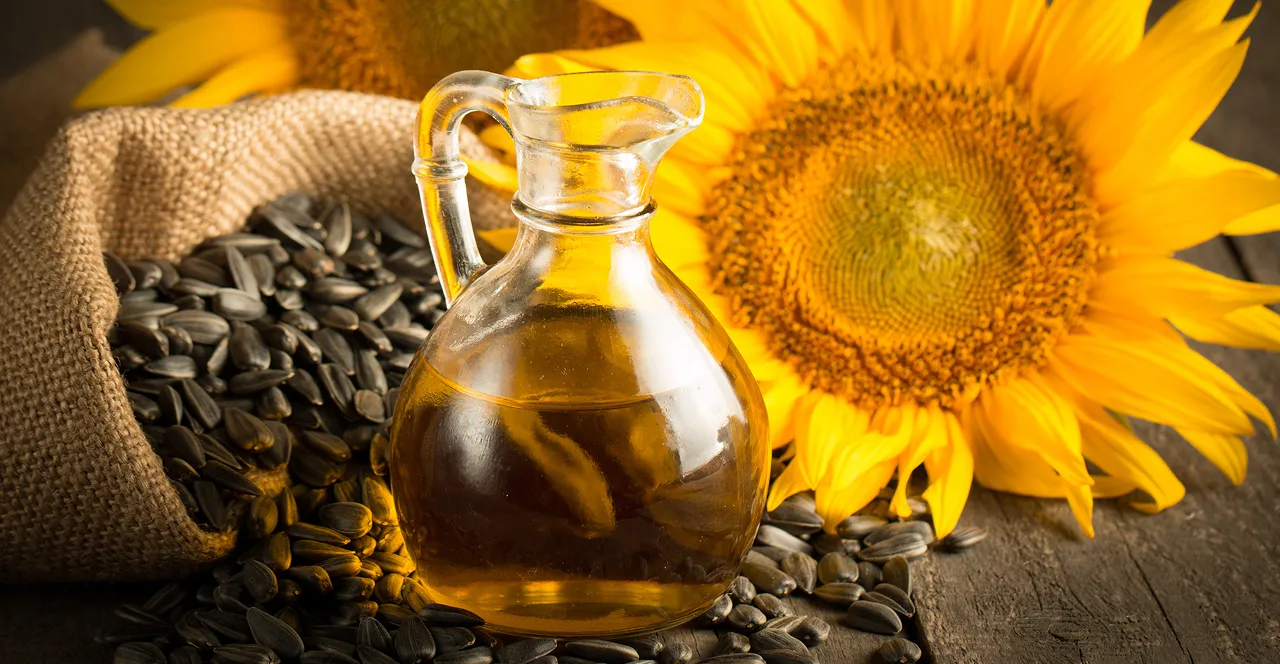 Glass jug of sunflower oil surrounded by sunflower seeds spilling from a burlap sack and a large bright sunflower on a wooden surface.