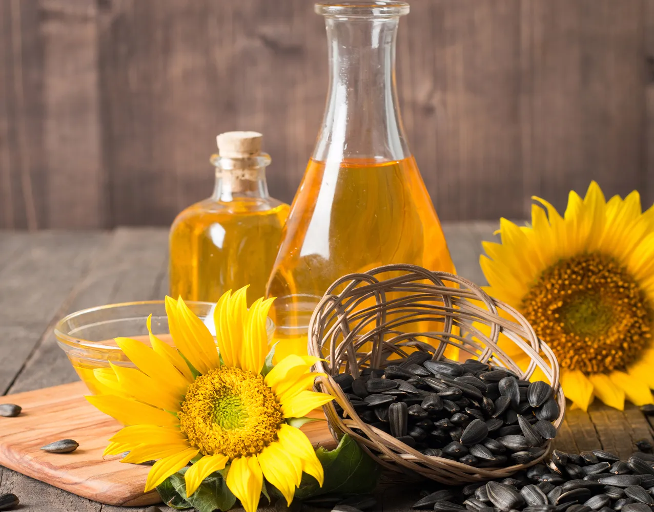 Sunflower blossoms, a wicker basket filled with black sunflower seeds, and bottles of sunflower oil on a wooden surface.