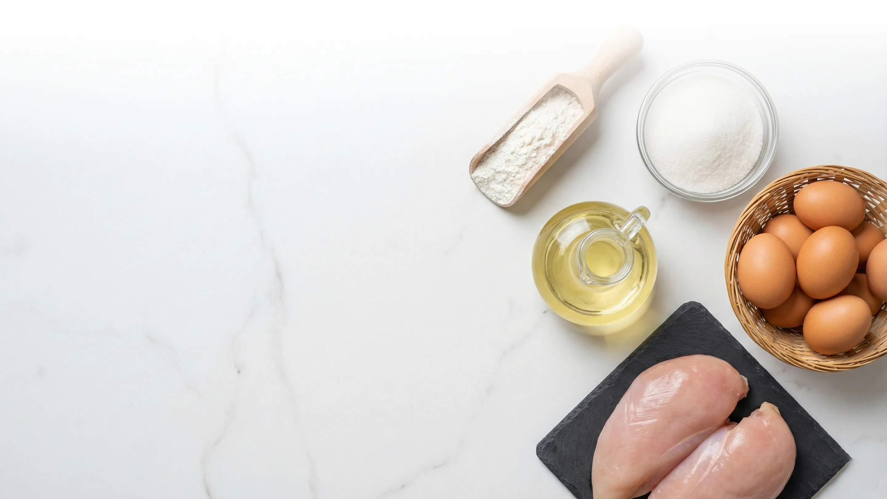 Raw chicken breasts on a black slate board with a basket of brown eggs, a glass bowl of sugar, a wooden scoop of flour, and a glass pitcher of oil on a white marble surface.
