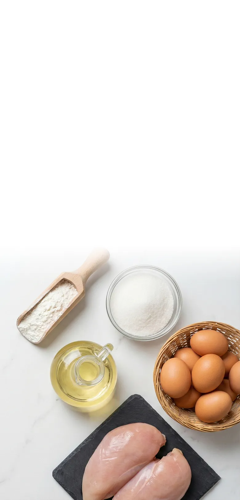 Raw chicken breasts on a black slate next to a basket of brown eggs, a glass jar of oil, a bowl of sugar, and a wooden scoop of flour on a white surface.