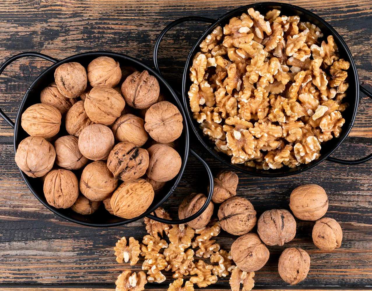 Two black bowls on wood table, one with whole walnuts, the other with shelled walnut halves. Armin Plus