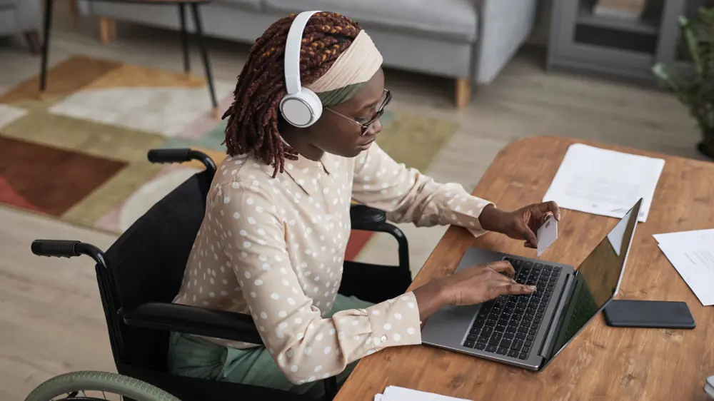Barrierefreiheit im Büro: Frau im Rollstuhl arbeitet konzentriert am Laptop. Moderne Arbeitsumgebung inklusiver Arbeitsplatz für Menschen mit Behinderungen, digitales Arbeiten, Online-Kommunikation, Homeoffice-Ausstattung, Diversity am Arbeitsplatz.
