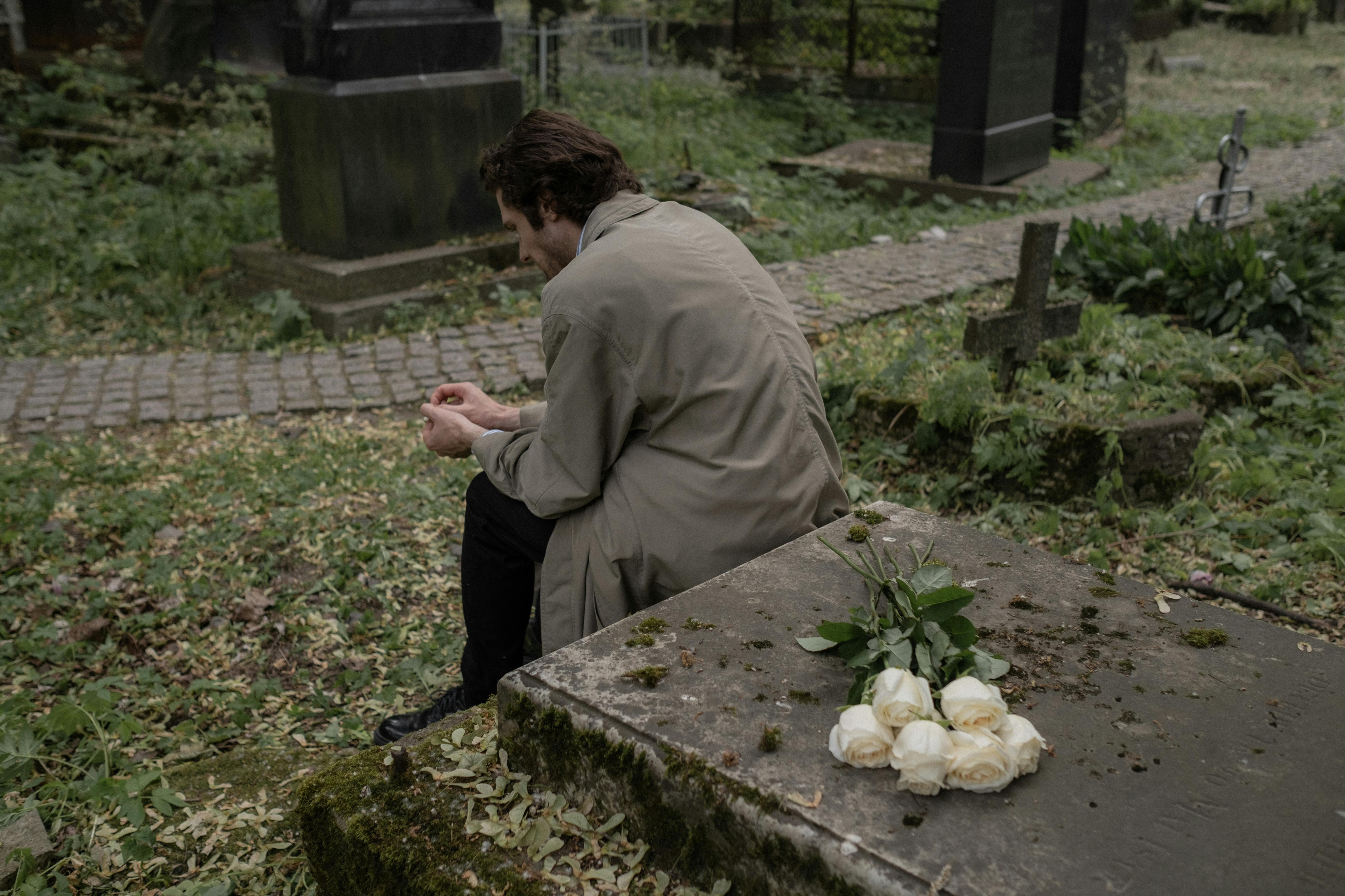 A man sits on a tombstone surrounded by flowers, reflecting on loss and resilience in support of grieving families.