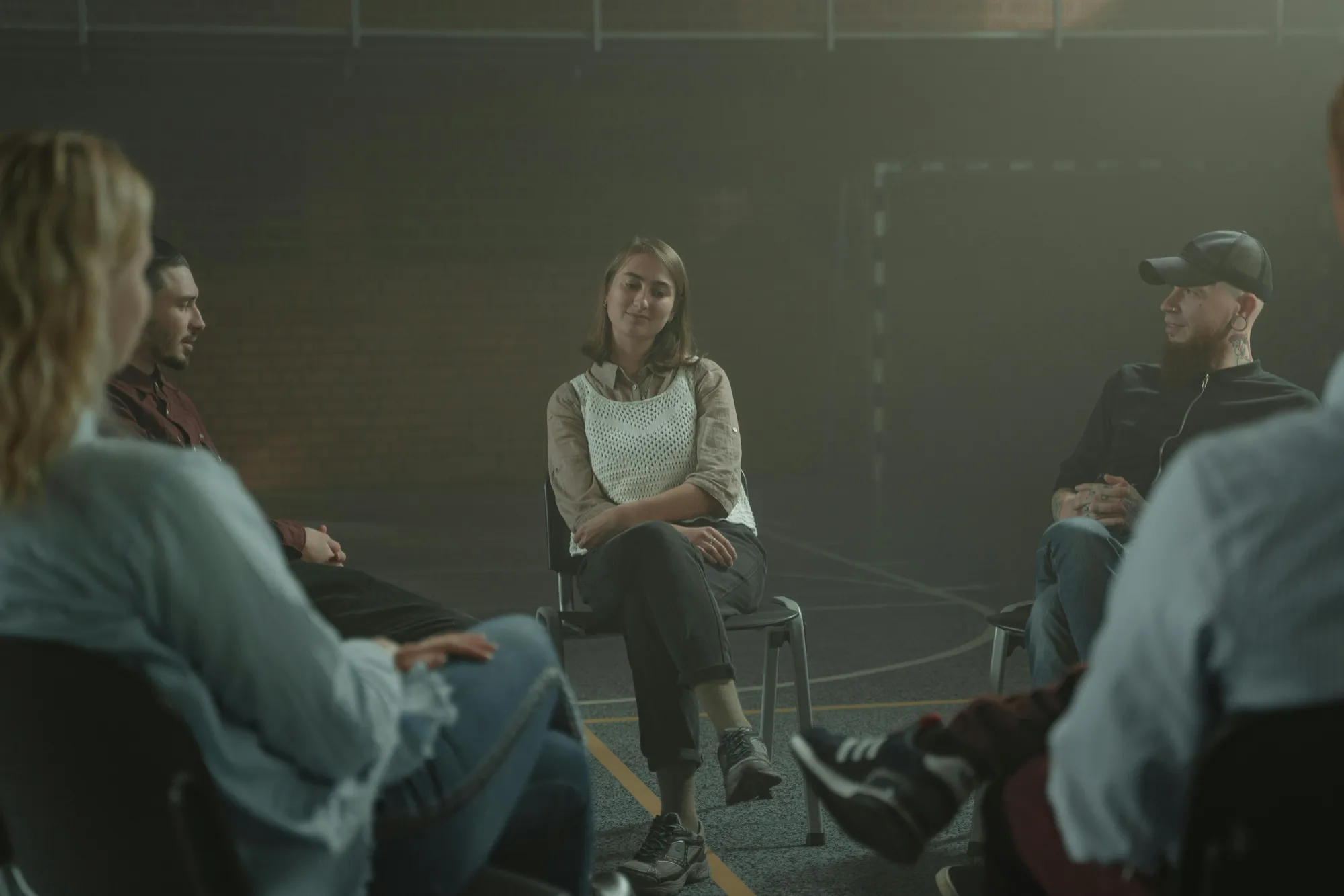 A group of people sit in a circle, engaged in a calm, introspective discussion. The setting is dimly lit, creating a reflective and supportive atmosphere.