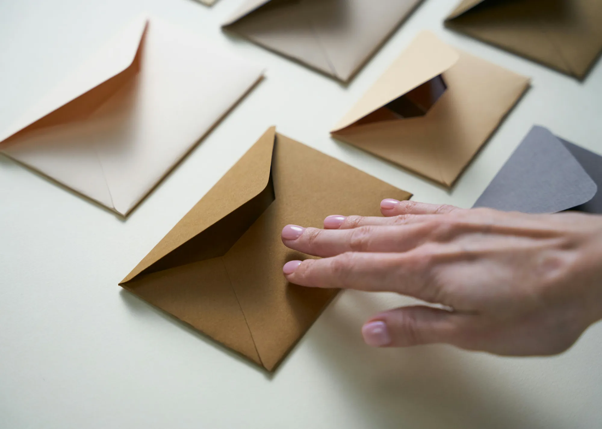 A hand gently touches a tan envelope, part of an arrangement of open and closed envelopes in shades of brown on a light surface, evoking curiosity.