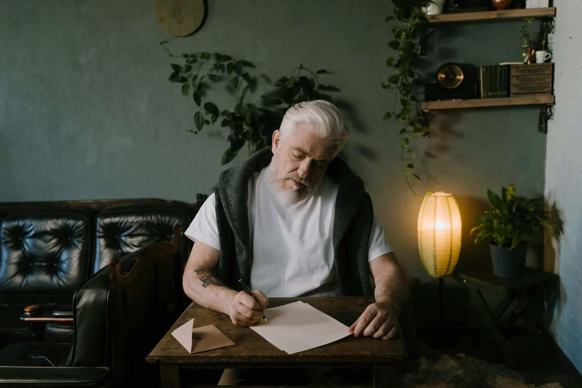 Elderly man writing at a wooden desk in a cozy room, with warm lighting and plants. He appears focused and calm. Leather sofa in the background.