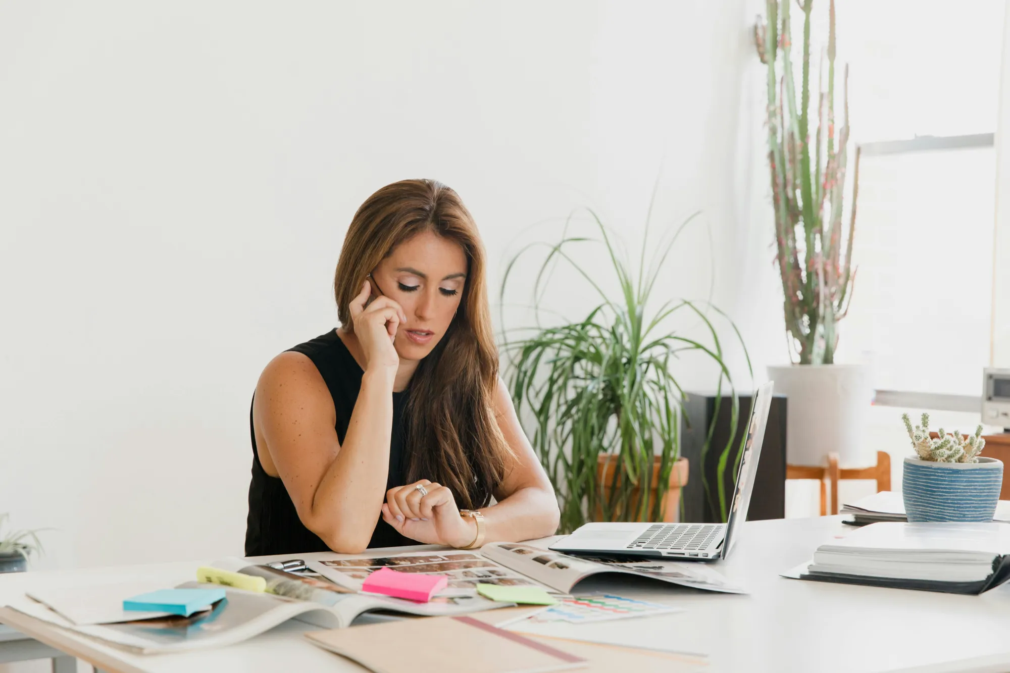 A woman sits at a white desk, looking thoughtful and focused, talking on the phone. Open magazines, sticky notes, and a laptop are in front of her, with tall plants in the background.