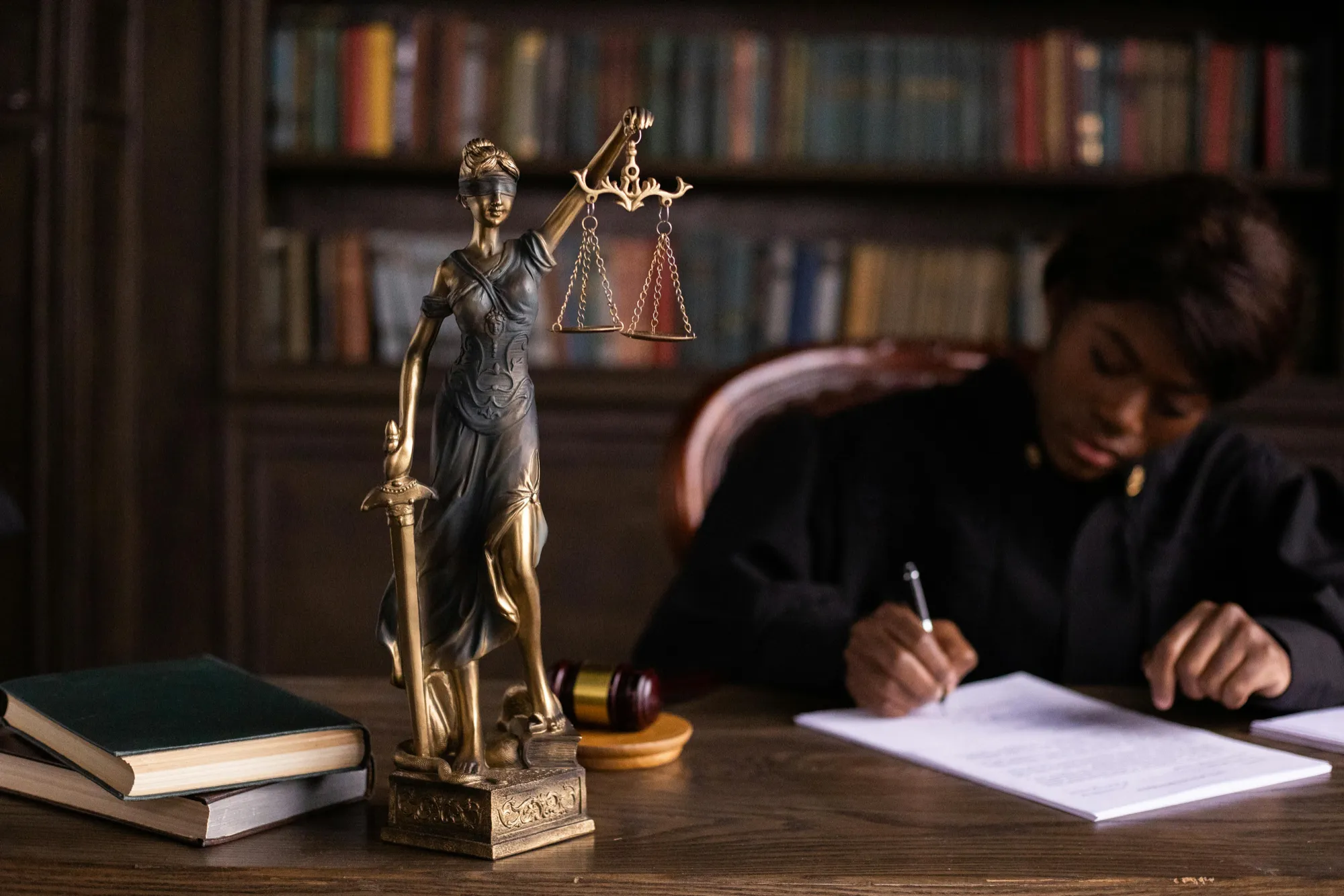 A brass Lady Justice statue stands prominently on a desk with scales in one hand, surrounded by books and a gavel. In the background, a person in a dark robe writes at the desk, with bookshelves behind them, creating a serious, scholarly atmosphere.