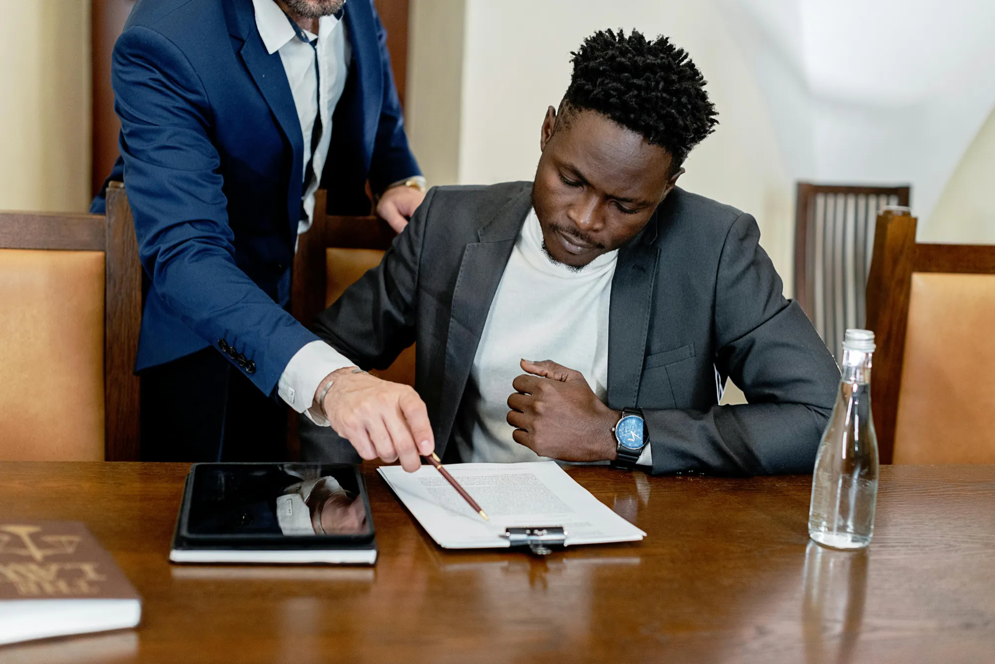 Man in a suit points at a clipboard document, guiding another man who is seated and focused on reading. A tablet and water bottle are on the table.