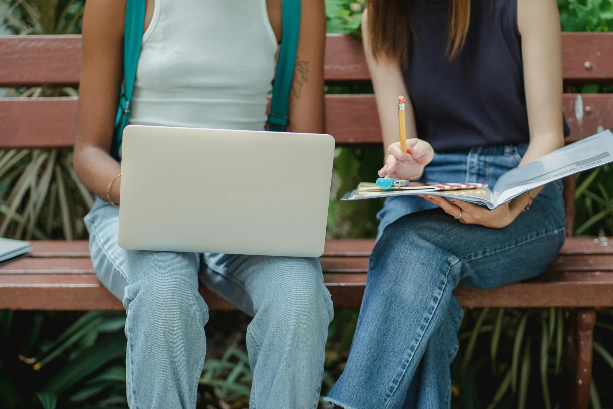 Two people sit on a park bench; one uses a laptop, the other writes in a notebook. Casual attire, green foliage in the background, study-focused mood.