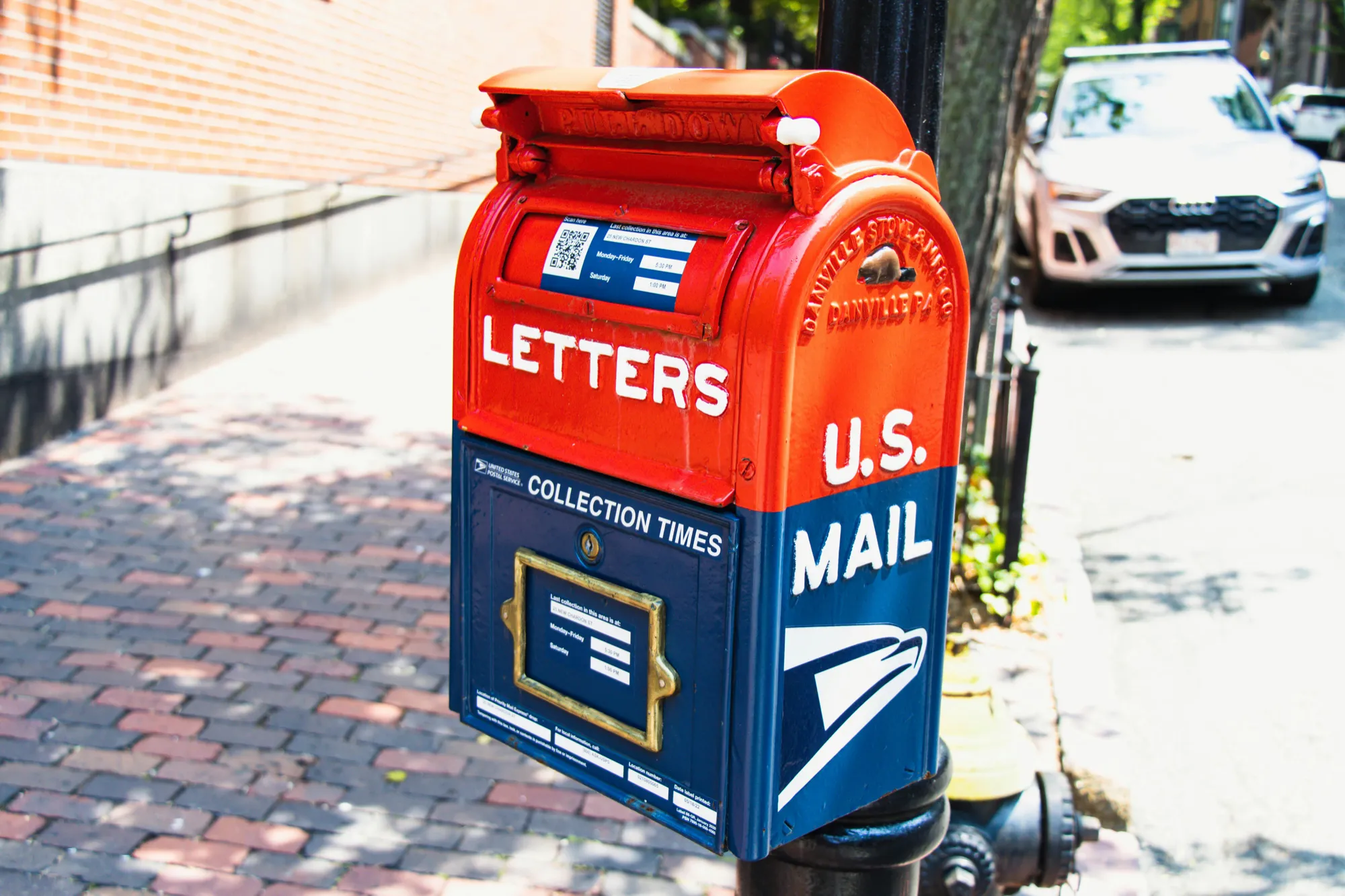 A red and blue U.S. Mail letterbox stands on a sunny street with brick pavement. A parked car is visible in the background, creating an urban atmosphere.
