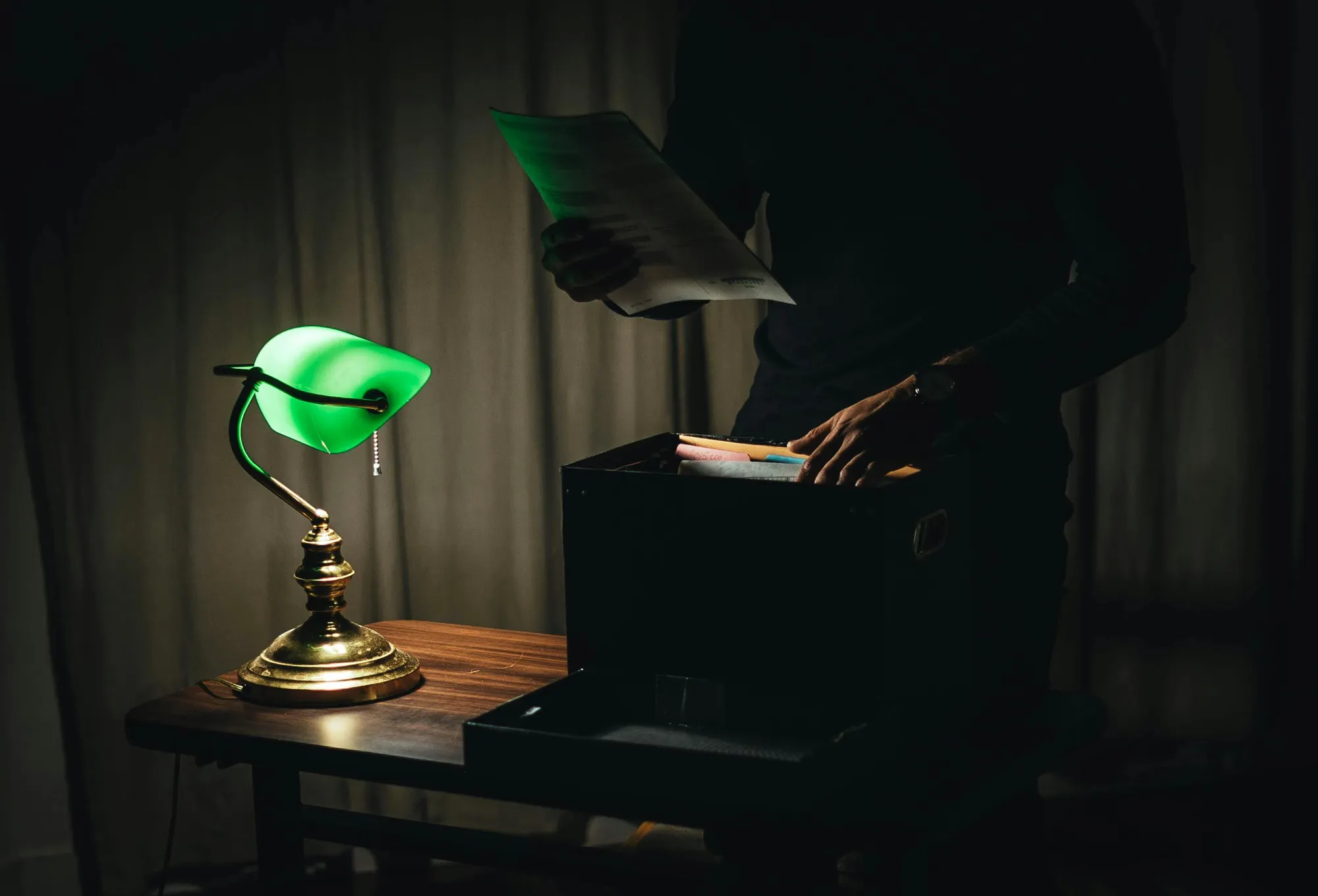 Dimly lit room with a green desk lamp illuminating a person holding a paper over an open file box on a wooden table, creating a mysterious ambiance.