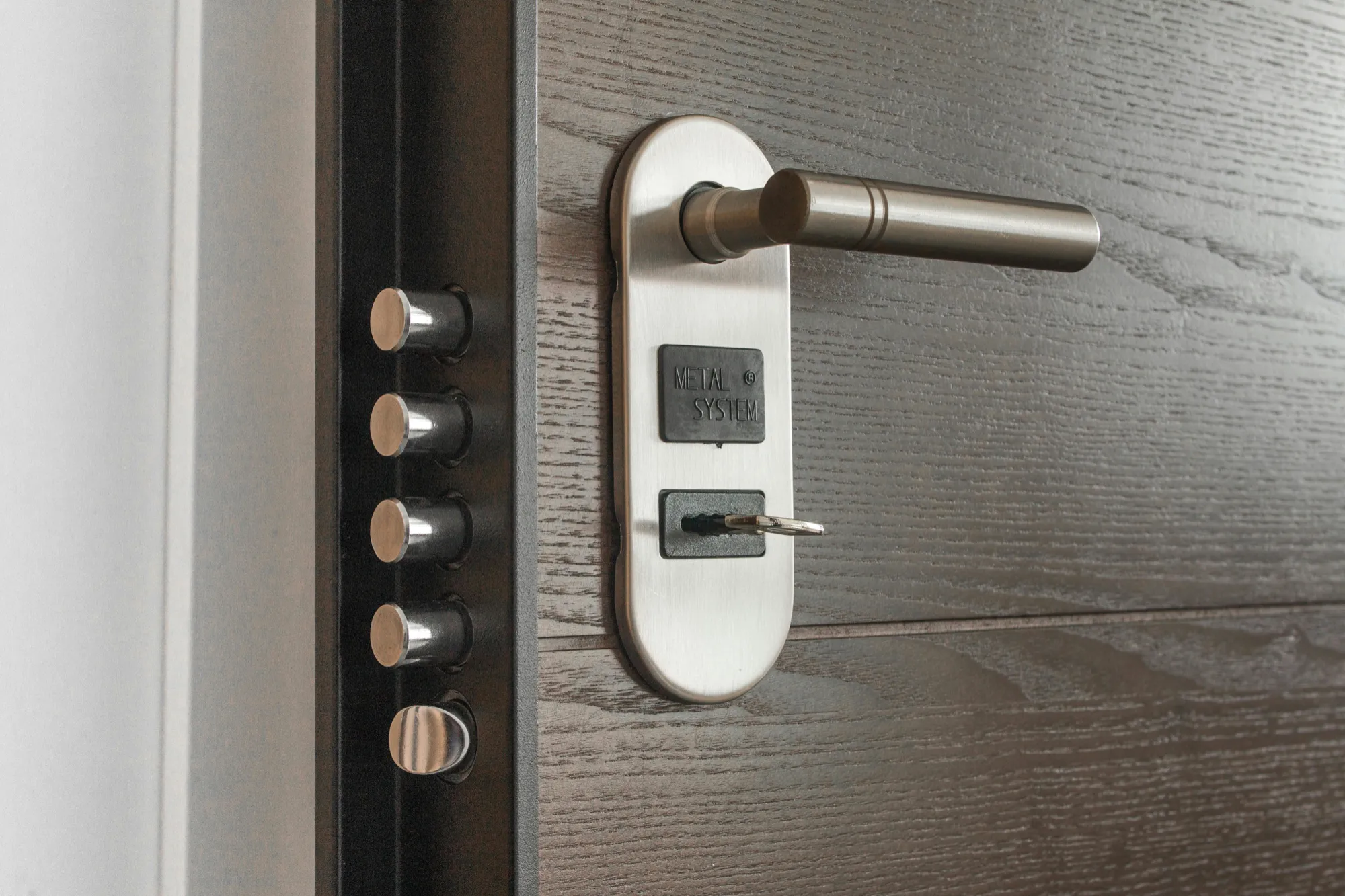 A close-up of a modern, wooden door with a sleek metal handle and key inserted in the lock. Four shiny bolts are visible on the door frame, emphasizing security.