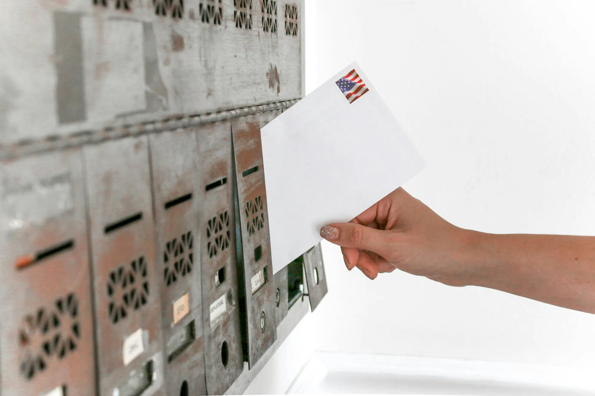 Person mailing a white envelope with an American flag stamp into a mailbox with multiple slots.