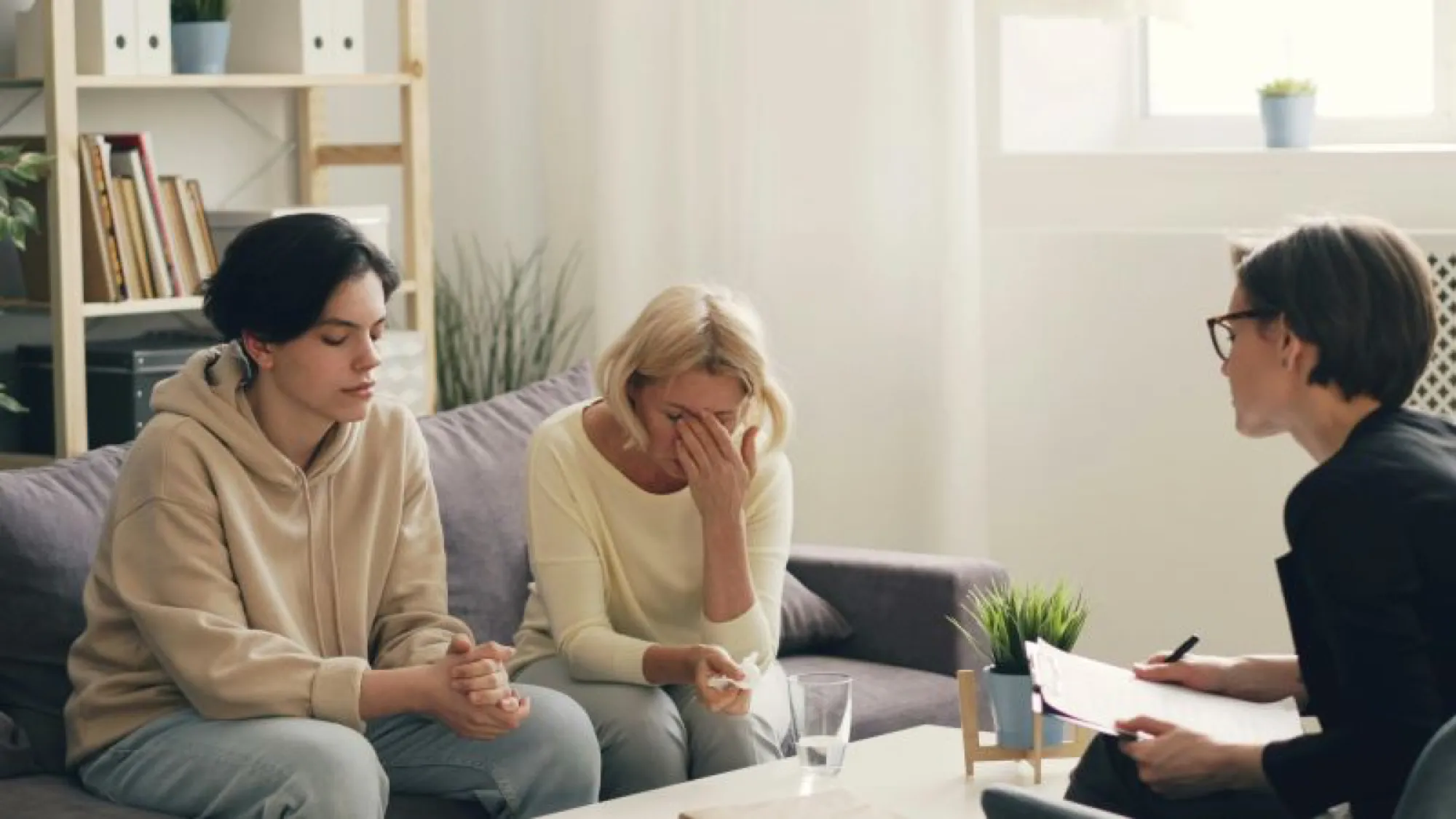 A woman speaks compassionately to a man and another woman on a couch, discussing grief and estate planning after a loss.