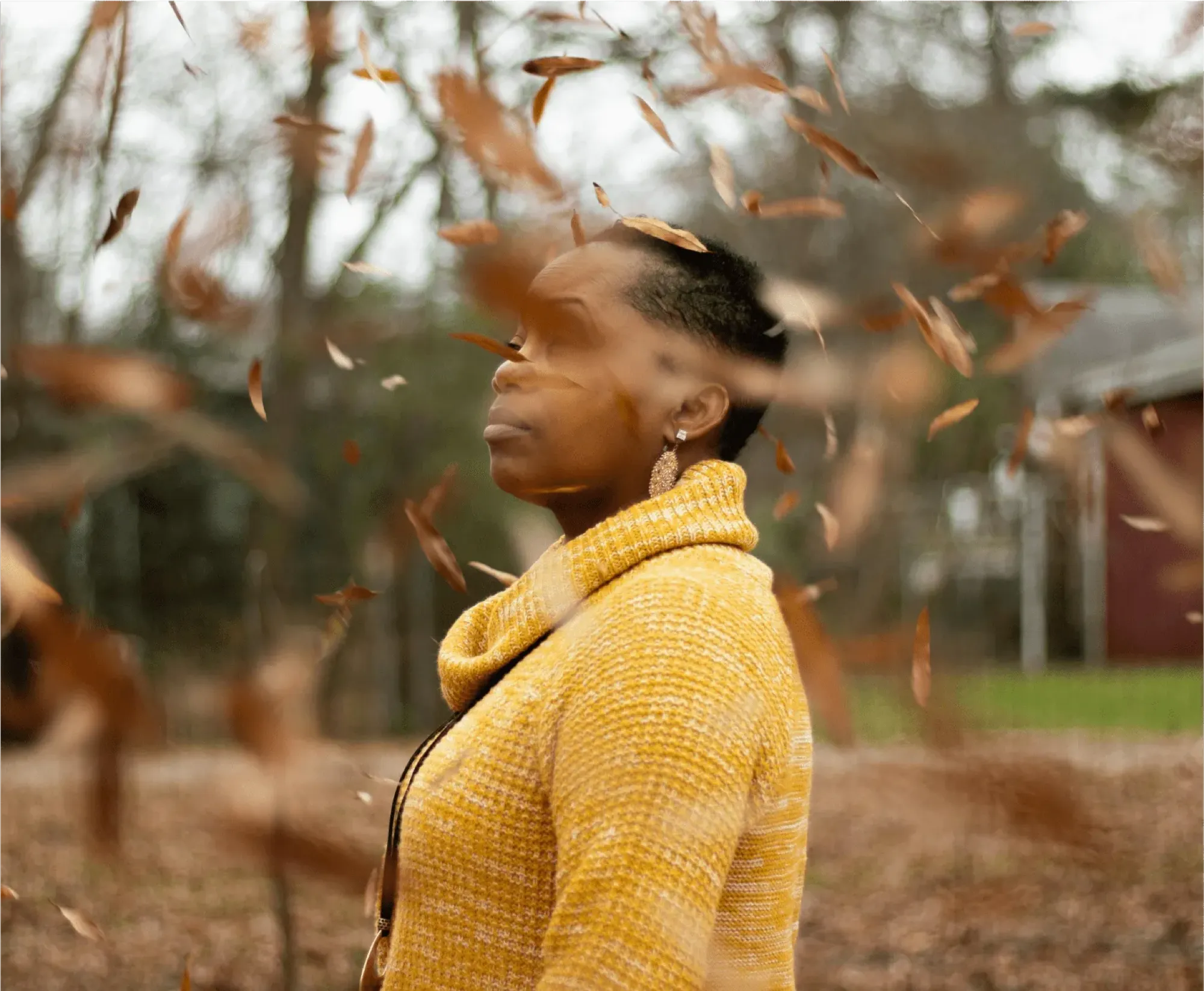 A woman in a yellow sweater stands amidst a swirl of colorful falling leaves in an autumn setting.