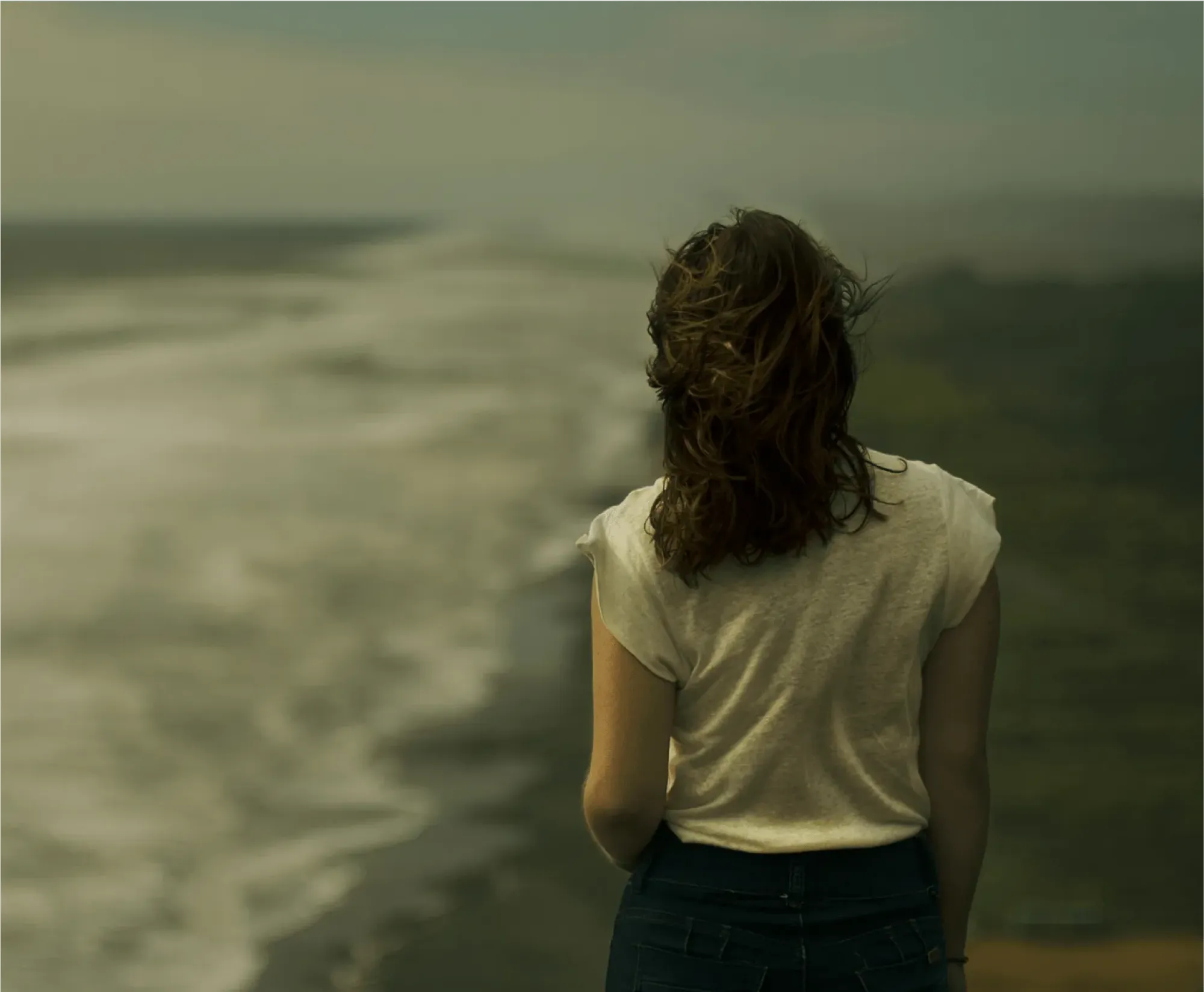 Woman in a white T-shirt seen from behind looking out over a misty coastline