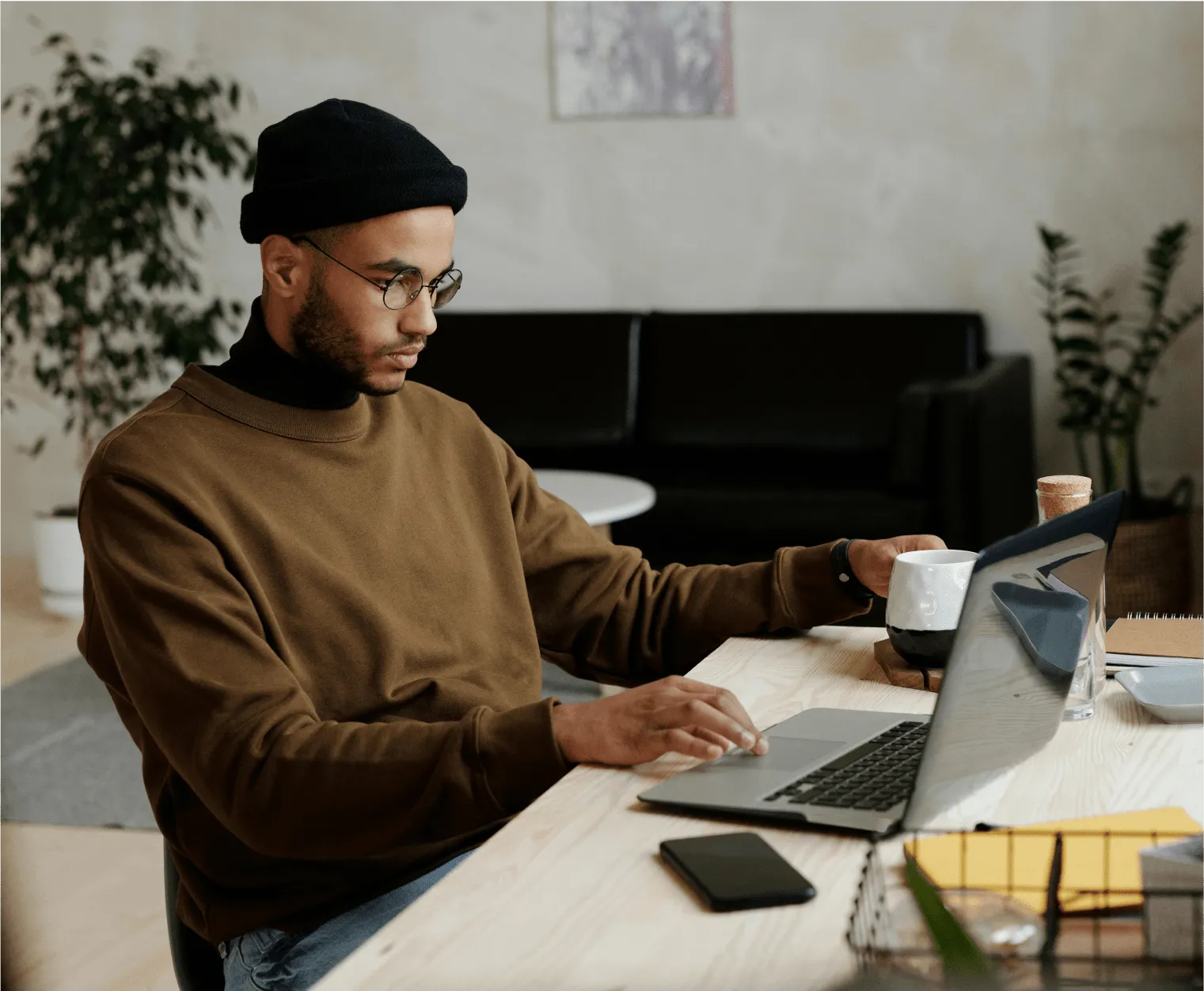 Man in a beanie and glasses working on a laptop at a wooden desk, holding a mug