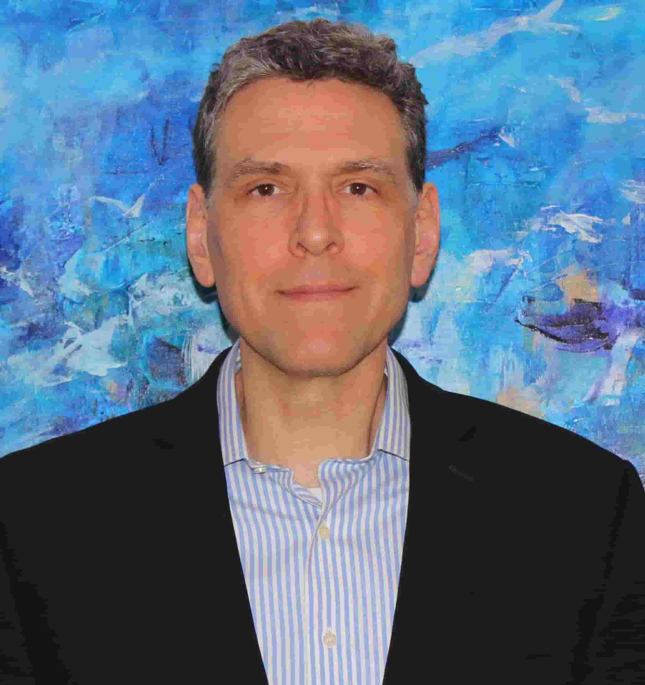 Woman with light brown hair wearing a white blouse sitting on a sofa with folded hands, smiling gently.