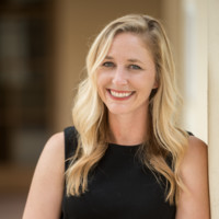 Woman with light brown hair wearing a white blouse sitting on a sofa with folded hands, smiling gently.