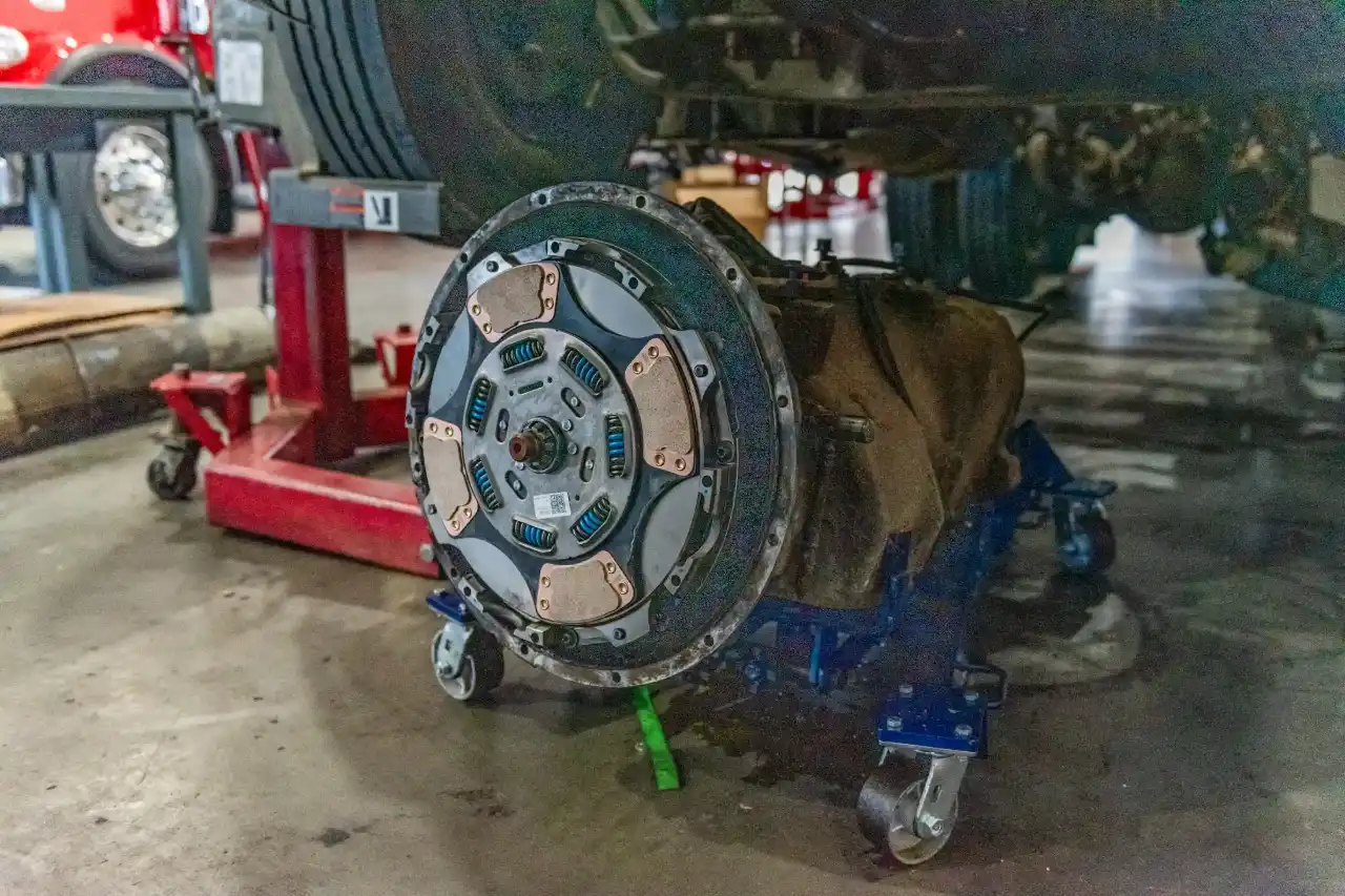 Flywheel and multi-puck clutch disc on floor jack under lifted truck, QR sticker visible, awaiting drivetrain reassembly.