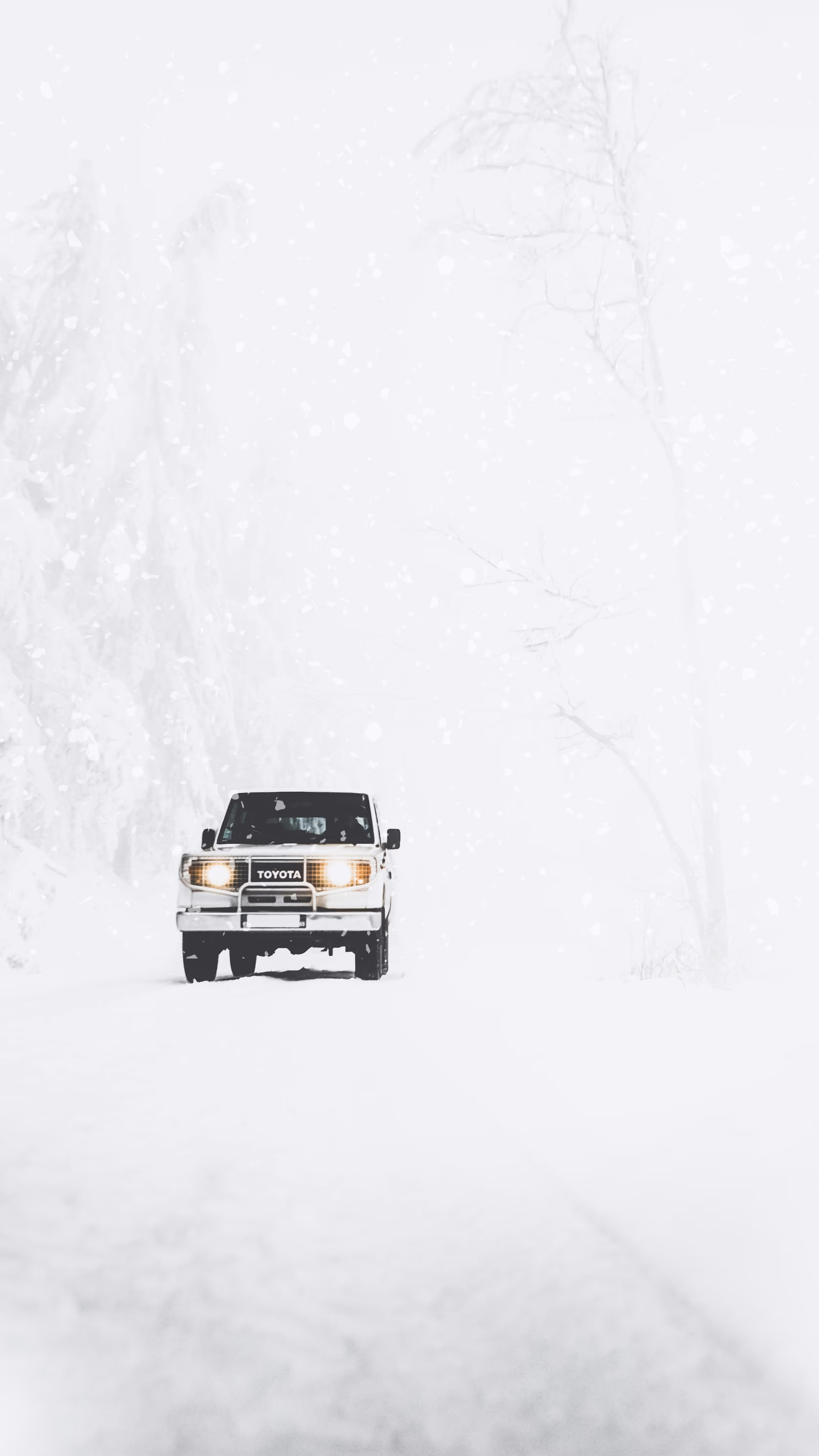 A lone white Toyota car on a snow-covered road amidst a whiteout, conveying the concept of winter travel or challenging driving conditions.