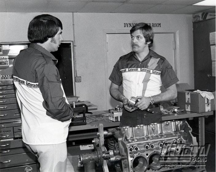 Robert Yates holding an engine component while speaking with a colleague in a dynamometer room