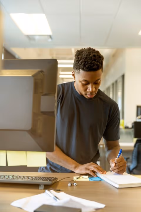 A remote assistant working at a standing desk, writing notes in a notebook while looking at a computer screen. The workspace includes a monitor, keyboard, sticky notes, and office supplies, representing essential skills for remote assistants such as time management, organization, and technological proficiency.